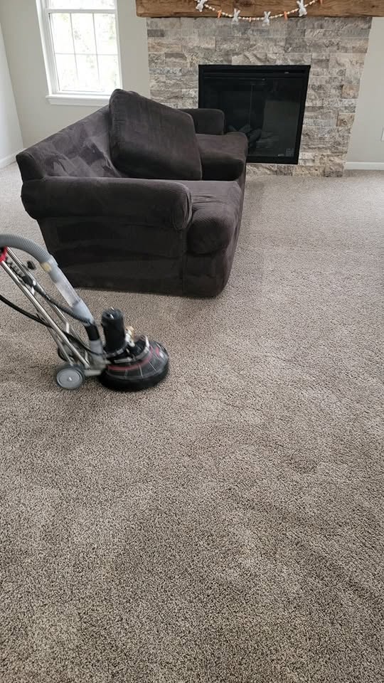 A professional carpet cleaning machine agitates and cleans a taupe residential carpet in a living room with a dark brown sofa and stone fireplace in the background, showing a distinct clean line.